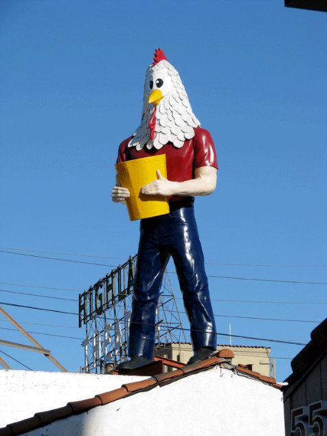 A large statue of a humanoid chicken wearing jeans, a yellow t-shirt and holding a bucket of fried chicken.