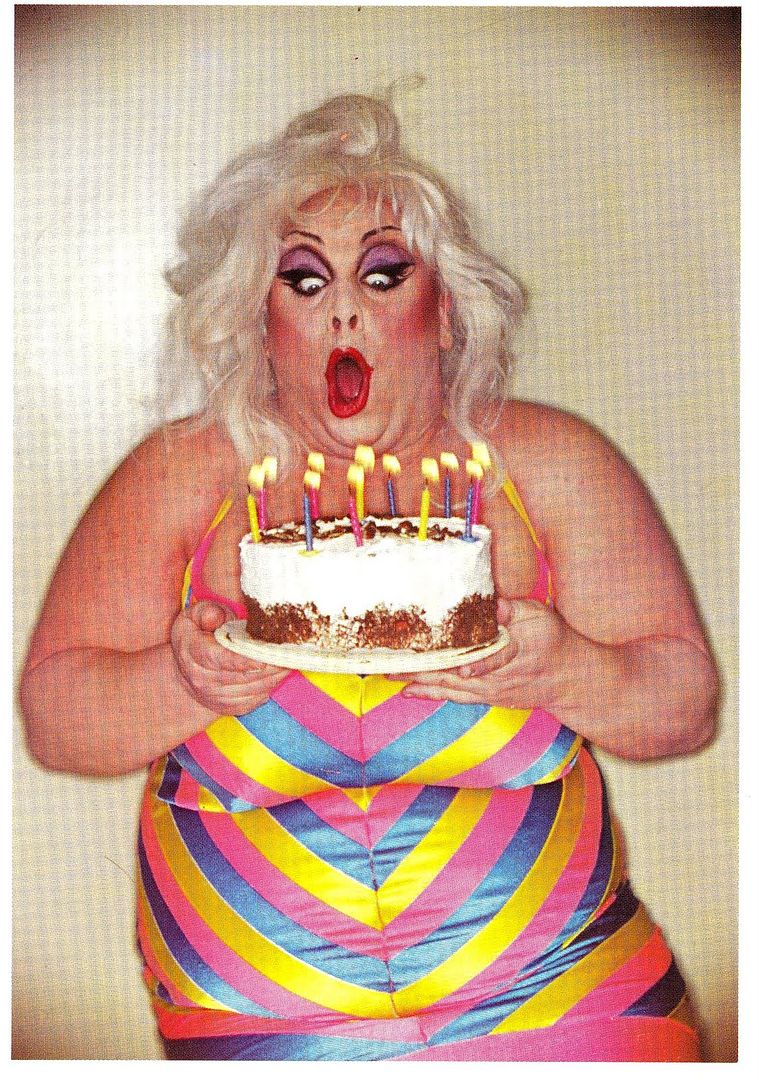 Divine, holding a birthday cake and blowing out the candles.